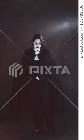 Man in black suit and cape, as vampire, faces forward, displaying fangs, in dimly lit, smoky environment. Lord of darkness. Man in black suit and cape, as vampire, faces forward, displaying fangs, in dimly lit, smoky environment. Lord of darkness. 121788336