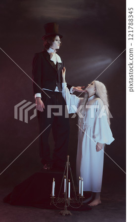 Man in black suit and top hat, looks as vampire, stands elevated, while woman in white reaches toward him, with lit candles at bottom of frame. Man in black suit and top hat, looks as vampire, stands elevated, while woman in white reaches toward him, with lit candles at bottom of frame. 121788345