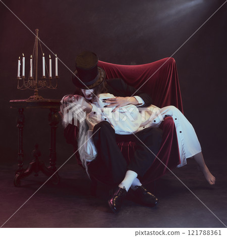 Man, vampire, in dark suit with cape leans over woman in white clothing, holding her as he gazes with sharp intensity. 121788361