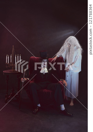 Terrified man in dark clothing, vampire, sits on chair, accompanied by woman in white gown standing beside him, both under soft dramatic light. 121788364