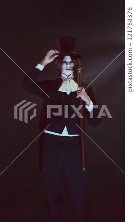 Pale-faced man, vampire, dressed in sleek black attire and top hat stands holding cane, looking serious under dim light. 121788378