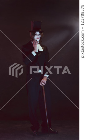 Pale-faced man, vampire, dressed in sleek black suit and top hat stands holding cane, looking serious under dim light. 121788379