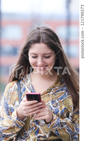 Beautiful woman with long blond hair holding mobile phone, looking at mobile phone screen, using smartphone apps. posing on street. Positive model smiling. She is standing with smartphone in hands on 121790273