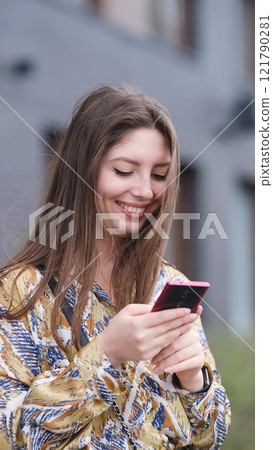 Beautiful woman with long blond hair holding mobile phone, looking at mobile phone screen, using smartphone apps. posing on street. Positive model smiling. She is standing with smartphone in hands on 121790281