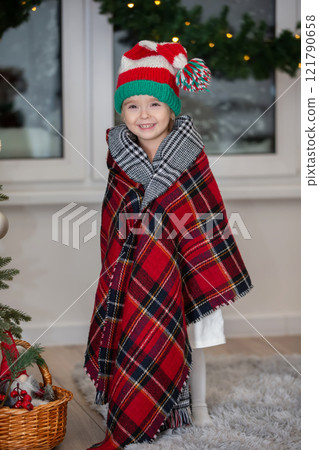 Happy little girl wearing santa claus hat wrapped in warm checkered blanket in christmas room. 121790658