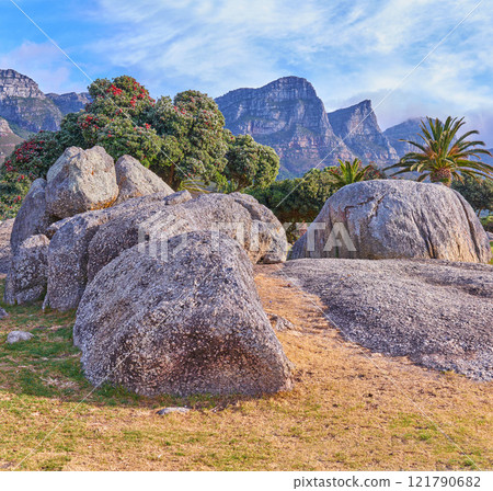 Rocks and boulders against a majestic mountain background with lush green trees and a clear blue sky. Remote and quiet landscape on a cliff with stones to explore during a scenic hiking in summer 121790682