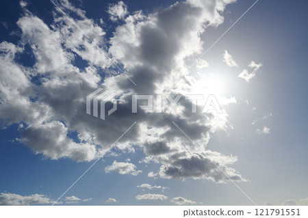 Clouds. Sun hiding behind clouds with blue sky. Beautiful sunshine from behind white and grey cumulus clouds at midday in summer. Serene view of natures outdoor beauty high in space. 121791551