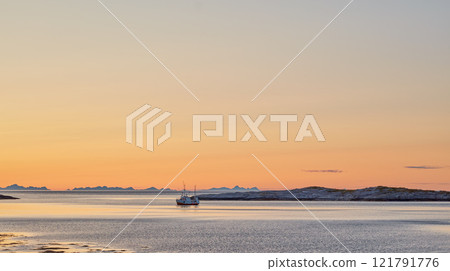 Distant sailboat in the sea near a mountain in the water during sunset. The ocean, beach, or large lake with a marine vessel in the distance near a hill outdoors during dawn on a summer day in Norway 121791776