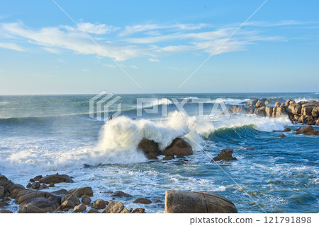A rocky coastline in the Cape Province, South Africa. Ocean waves crashing on coastal rocks on a sunny summer day with blue clear skies and a scenic tropical landscape beachfront in the Western Cape 121791898
