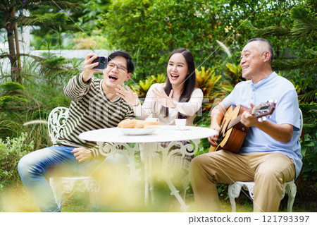 Happy multi-generation Asian family living together in weekend. Happy Asian family spending time together, man and woman let their father playing a guitar and singing in backyard garden at home.  121793397