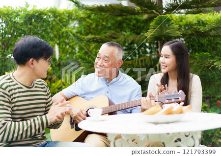 Happy multi-generation Asian family living together in weekend. Happy Asian family spending time together, man and woman let their father playing a guitar and singing in backyard garden at home.  121793399