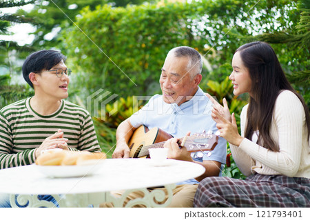 Happy multi-generation Asian family living together in weekend. Happy Asian family spending time together, man and woman let their father playing a guitar and singing in backyard garden at home.  121793401