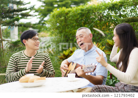 Happy multi-generation Asian family living together in weekend. Happy Asian family spending time together, man and woman let their father playing a guitar and singing in backyard garden at home.  121793402