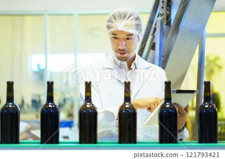 An empty glass bottles are rolling on the alcohol bottling belt machine in the manufacturing facility, meanwhile, male quality control supervisor is examining a bottles on the line. An empty glass bottles are rolling on the alcohol bottling belt machine in the manufacturing facility, meanwhile, male quality control supervisor is examining a bottles on the line. 121793421