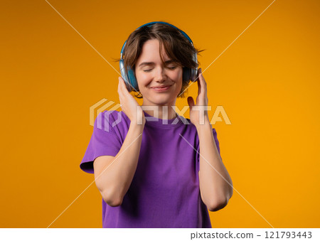 Joyful woman listening music in vibrant yellow studio, headphones on, fully immersed in. Her energy and happiness fill room as she moves with confidence and rhythm. 121793443