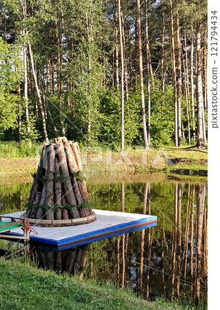 Traditional bonfire structure built with logs and greenery, placed on floating platform in serene forest setting by a calm lake 121794434