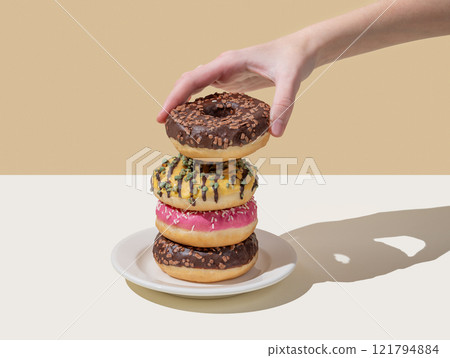 Hand takes one donut from stack of glazed donuts with different toppings on beige white background. Hand takes one donut from stack of glazed donuts with different toppings on beige white background. 121794884
