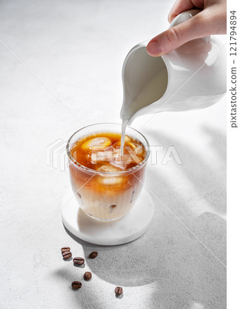 Cold latte coffee in a glass with hand pouring alternative milk from a milk jug on light background 121794894