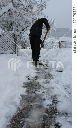 man from behind in black clothes clearing path between snowdrifts in backyard or garden after snow storm, seasonal winter work in rural area, cleaning snow with shovel after snowfall man from behind in black clothes clearing path between snowdrifts in backyard or garden after snow storm, seasonal winter work in rural area, cleaning snow with shovel after snowfall 121795284