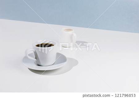 Closeup of cup with black coffee on a saucer and creamer on white table with blue background 121796853