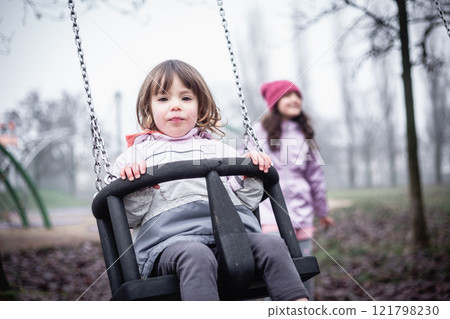 one small child sitting on the swing at children playground on a foggy day, playing with other child 121798230
