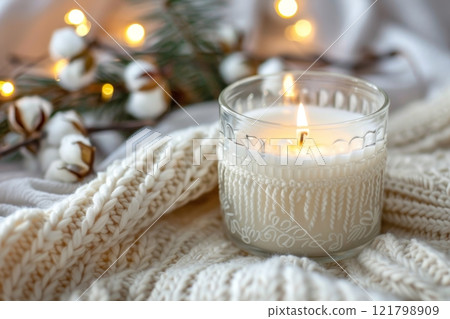 A beautifully styled candle in a transparent glass container emits a warm glow, set among soft textures and twinkling lights 121798909