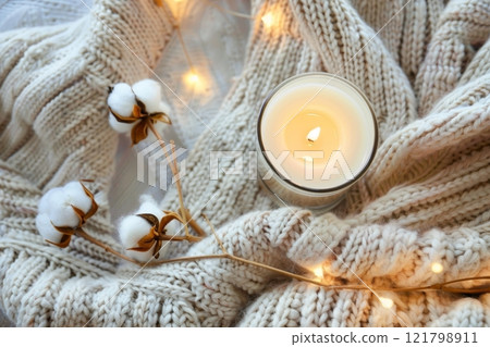 A warm candle is placed in a glass vessel, nestled within a soft knit blanket and accompanied by decorative cotton stems and fairy lights A warm candle is placed in a glass vessel, nestled within a soft knit blanket and accompanied by decorative cotton stems and fairy lights 121798911