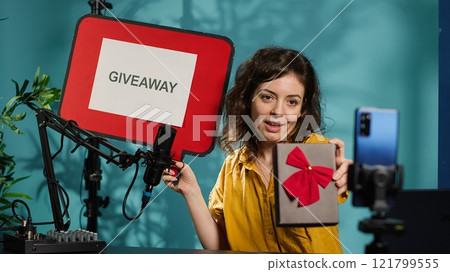 Smiling woman holding empty copy space sign, excited to do giveaway for subscribers. Media star happy to offer packed gift with ribbon to viewers, holding cardboard bubble speech cutout, camera A 121799555
