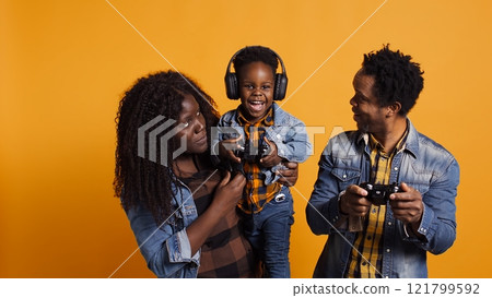 Joyful optimistic dad playing online video games with his boy using controller, parents enjoying leisure time with their small child. African american happy family play together in studio. Camera B. 121799592