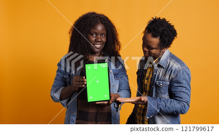 African american couple pointing at a tablet with isolated green screen display, smiling at each other against yellow background. Cheerful affectionate husband and wife posing. Camera B. African american couple pointing at a tablet with isolated green screen display, smiling at each other against yellow background. Cheerful affectionate husband and wife posing. Camera B. 121799617
