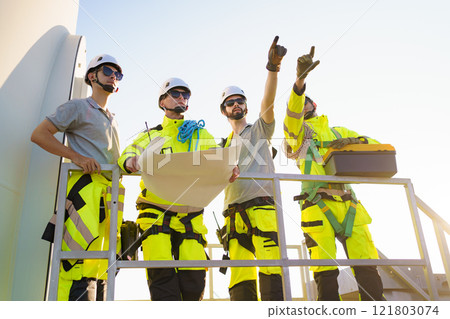 Team of professional electrical engineers in fully safety suit are working and discussing together at the windmill electric generating turbine. Electricians working at the site of windmill turbine. Team of professional electrical engineers in fully safety suit are working and discussing together at the windmill electric generating turbine. Electricians working at the site of windmill turbine. 121803074