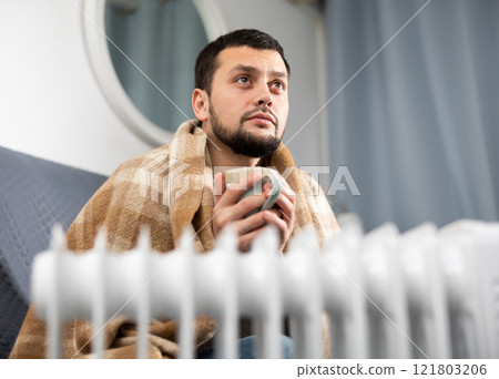 Young man wrapped in plaid sitting on sofa near radiator with cup Young man wrapped in plaid sitting on sofa near radiator with cup 121803206