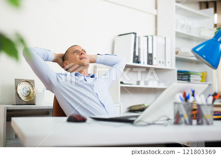 Relaxed office worker sitting at workplace with hands behind head Relaxed office worker sitting at workplace with hands behind head 121803369