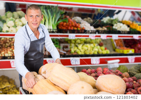 Smiling greengrocery owner laying out fruits and vegetables on counter 121803402
