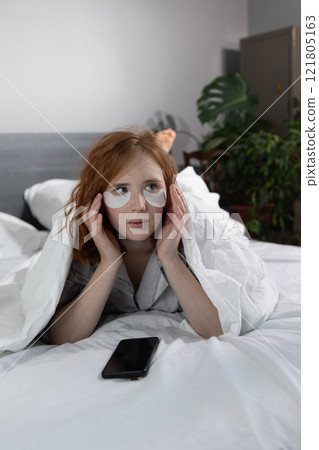 A woman lies comfortably in bed with eye patches, engrossed in her phone, enjoying her morning 121805163