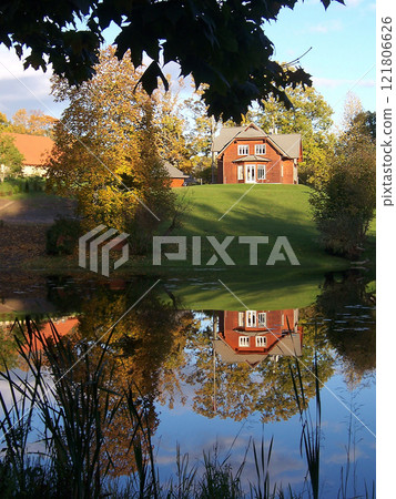 A small red house on a green hill is reflected in the mirror surface of the pond A small red house on a green hill is reflected in the mirror surface of the pond 121806626