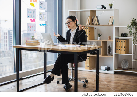 Asian woman in professional attire engaged in online video call from office desk. Business environment with laptop, headset modern, organized setting suggesting productivity and communication. 121808403
