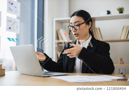 Asian businesswoman in headset conducting video call on laptop in modern office. Woman wearing glasses and suit appears professional and focused, engaging in conversation. Asian businesswoman in headset conducting video call on laptop in modern office. Woman wearing glasses and suit appears professional and focused, engaging in conversation. 121808404
