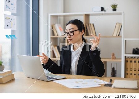 Asian businesswoman in black suit using laptop with headset during video call. Young professional engaging in online meeting in bright modern office setting with bookshelf and plant. 121808406