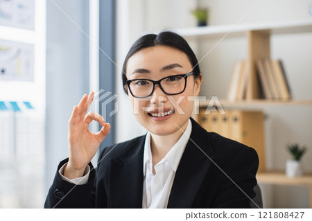 Asian businesswoman wearing glasses showing ok sign, smiling confidently in office setting. Captures positive energy, professional attire, and modern work environment, conveying success and approval. Asian businesswoman wearing glasses showing ok sign, smiling confidently in office setting. Captures positive energy, professional attire, and modern work environment, conveying success and approval. 121808427
