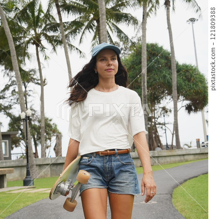 A woman is walking at the city park while carrying a skateboard in her hands A woman is walking at the city park while carrying a skateboard in her hands 121809388