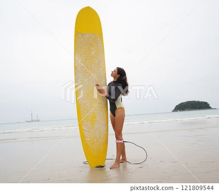 A woman in a stylish wet suit confidently holds a surfboard on the beach A woman in a stylish wet suit confidently holds a surfboard on the beach 121809544