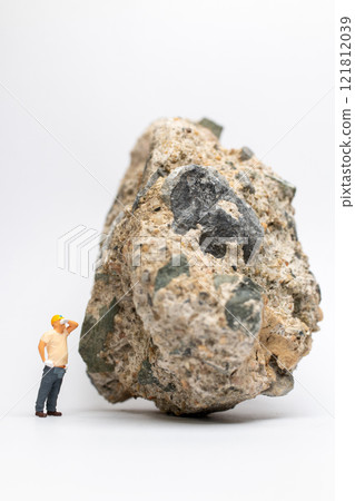 Miniature people construction worker stands next to a massive rock, scratching his head in contemplation Miniature people construction worker stands next to a massive rock, scratching his head in contemplation 121812039