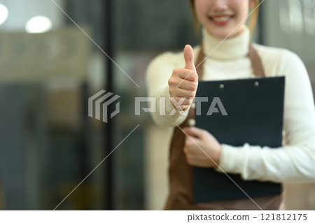 Cheerful coffee shop owner in an apron giving a thumbs up while holding a clipboard 121812175