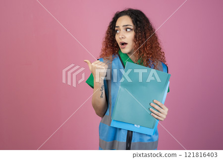 Front view of shocked, surprised woman with open mouth looking aside, pointing, holding folders. Isolated on pink studio background. Front view of shocked, surprised woman with open mouth looking aside, pointing, holding folders. Isolated on pink studio background. 121816403