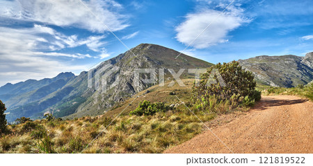 A hiking trail on a mountain with a cloudy blue sky on a summer day. A footpath surrounded by lush plants and grass outdoors on a spring afternoon. A pathway in nature or mountainside with copy space 121819522