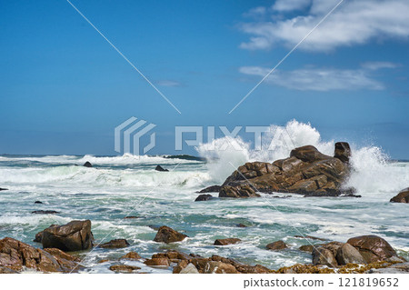 Big waves splashing and breaking on the coastline. Turbulent sea with rough tides from strong winds crashing onto beach boulders with a blue sky background. Rocky coast in Camps Bay, South Africa Big waves splashing and breaking on the coastline. Turbulent sea with rough tides from strong winds crashing onto beach boulders with a blue sky background. Rocky coast in Camps Bay, South Africa 121819652