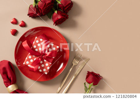 Valentine's day table setting with red plate, chocolate sweets, romantic gift and red roses on beige background. View from above. Valentine's day table setting with red plate, chocolate sweets, romantic gift and red roses on beige background. View from above. 121820804