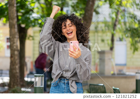 Caucasian woman use mobile smartphone celebrating win good message news outdoors on city street 121821252