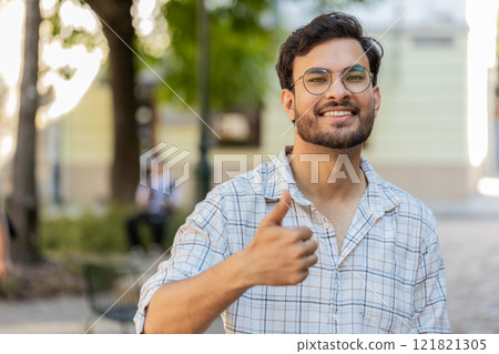 Happy young Indian man looking approvingly at camera showing thumbs up like gesture on city street Happy young Indian man looking approvingly at camera showing thumbs up like gesture on city street 121821305